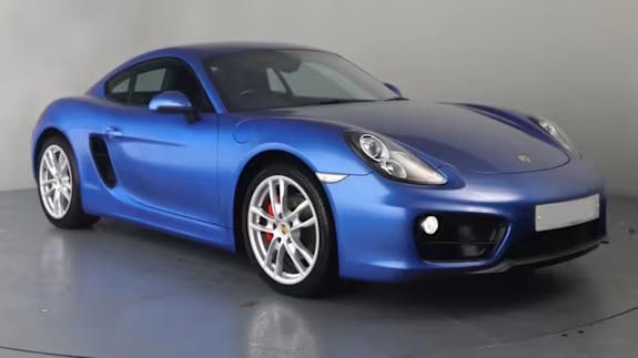 Matte blue Porsche Cayman coupe parked in a studio with a plain grey background, shown from the front three‑quarter view.