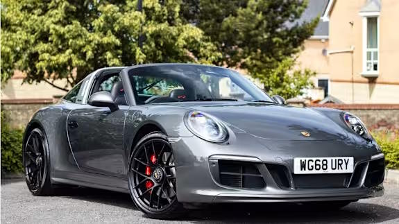 Grey Porsche 911 targa with black wheels and red brake calipers, parked on tarmac with trees and houses in the background, seen from the front three‑quarter view.