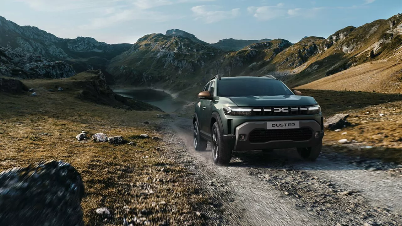 Dacia Duster driving along a rugged mountain trail, kicking up dust with rocky peaks and a lake in the background.