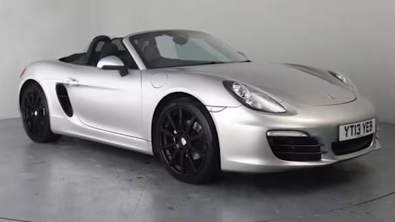 Silver Porsche Boxster convertible with black wheels, roof down, parked in a studio with a plain grey backdrop.