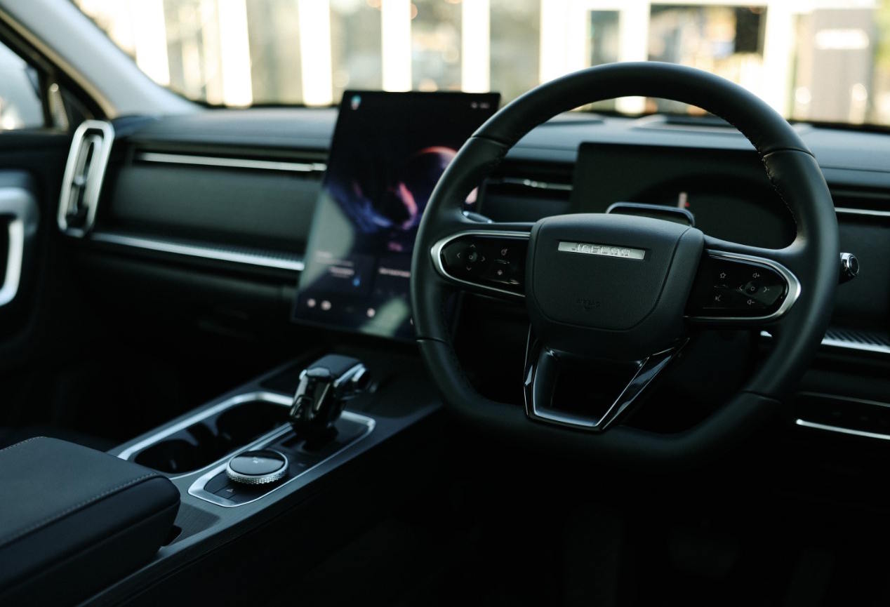 Interior view of the Jaecoo 7 SHS Hybrid, featuring a modern cockpit with leather-trimmed steering wheel, large portrait-style touchscreen, rotary gear selector and dark premium trim finishes.