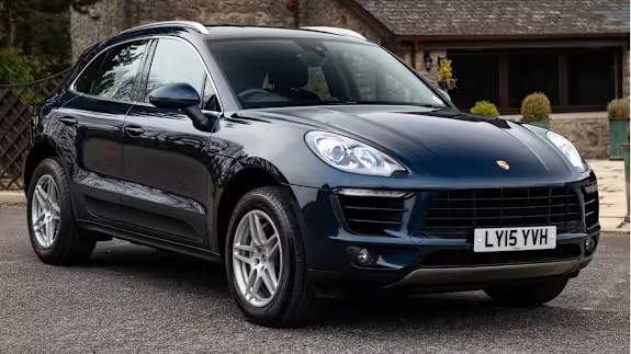 Dark blue Porsche Macan SUV parked on a driveway in front of stone buildings, viewed from the front three‑quarter angle.