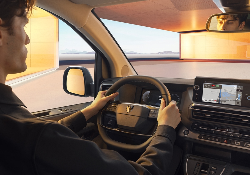 Driver holding the steering wheel inside Peugeot E-Traveller with digital display and navigation screen visible on the dashboard.