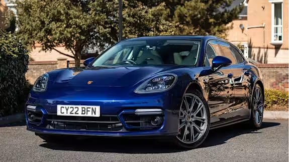 Dark blue Porsche Panamera Sport Turismo estate parked on tarmac in sunlight, with houses and trees behind, viewed from the front three‑quarter angle.