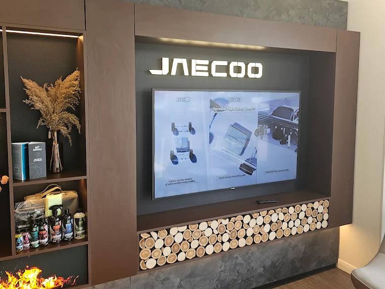 JAECOO Louth dealership showroom display featuring illuminated brand signage, interactive digital screen showcasing vehicle technology, and contemporary interior styling with decorative log feature