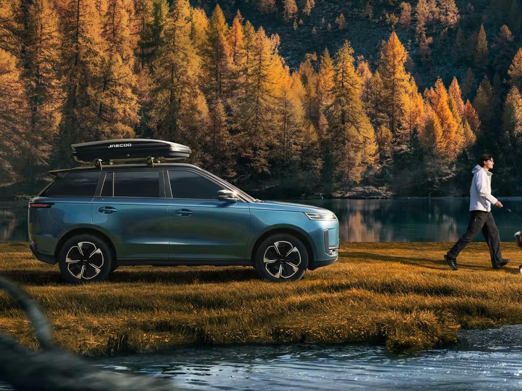 Side profile of the Jaecoo 5 SUV in teal blue, fitted with roof box and silver roof rails, parked lakeside against stunning autumn forest scenery.