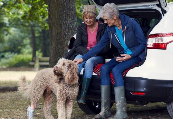 2 older women sat on the boot of a white car, leaning down to pet a Labradoodle.
