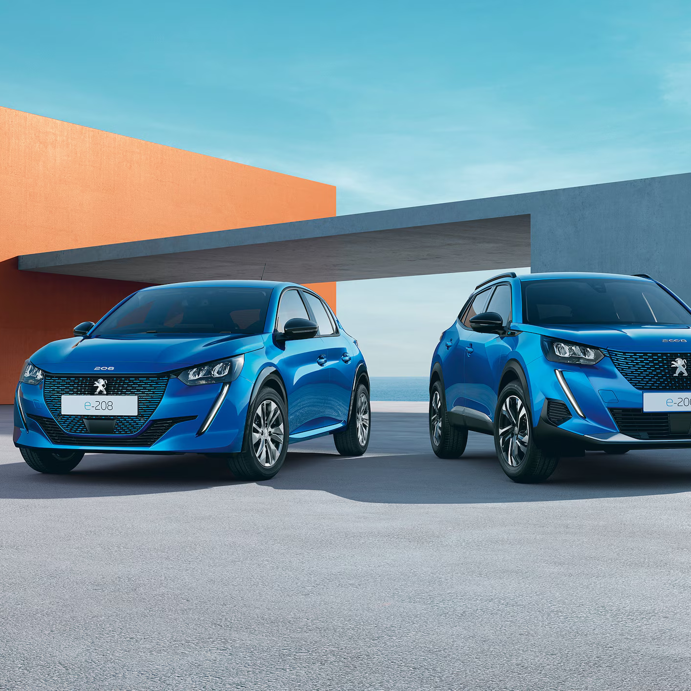 Two blue Peugeot e-208 cars parked in front of a modern architectural building with geometric shapes and a coastal backdrop.
