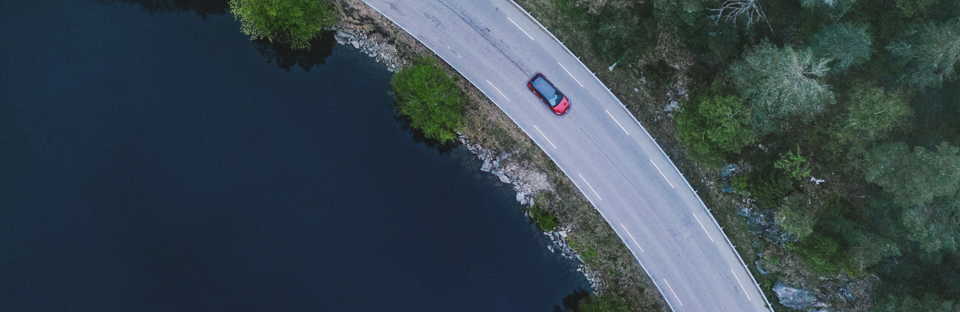 Ariel view of a red nissan car driving on a rural road next to a lake.