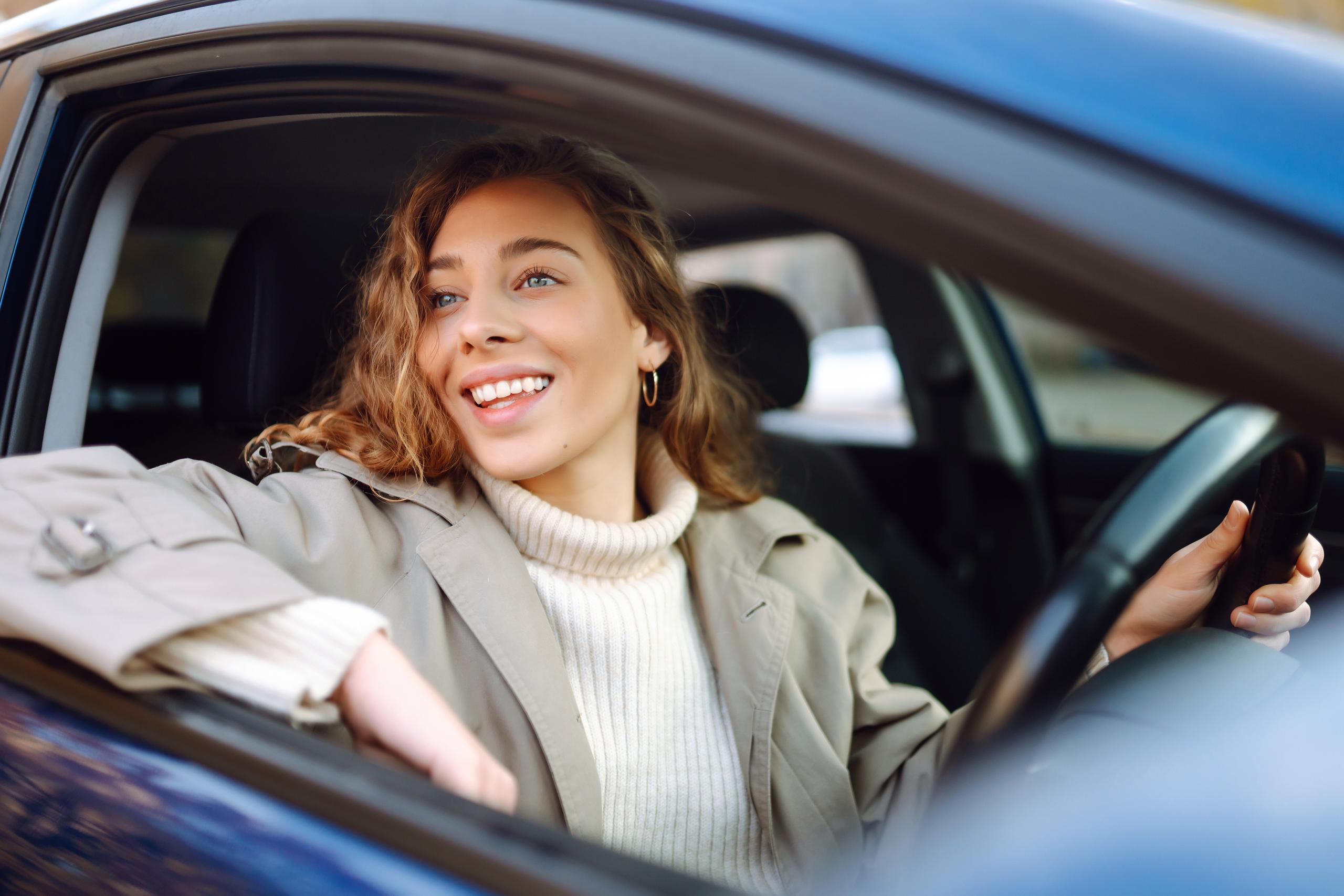 young woman driving a dark coloured car wearing a cream jumper and trench coat with one hand on the steering wheel