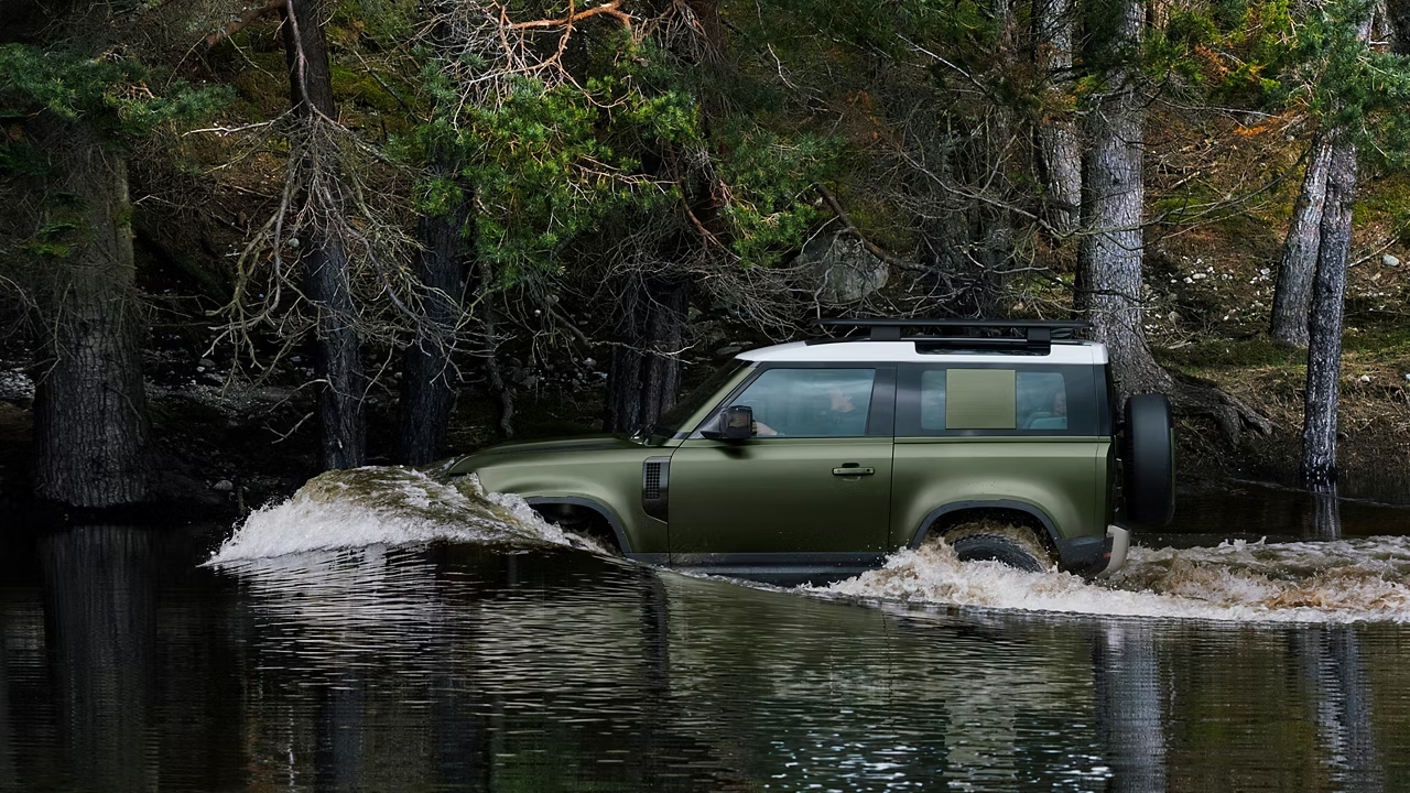 Green Defender 90 driving through shallow water in a forest.