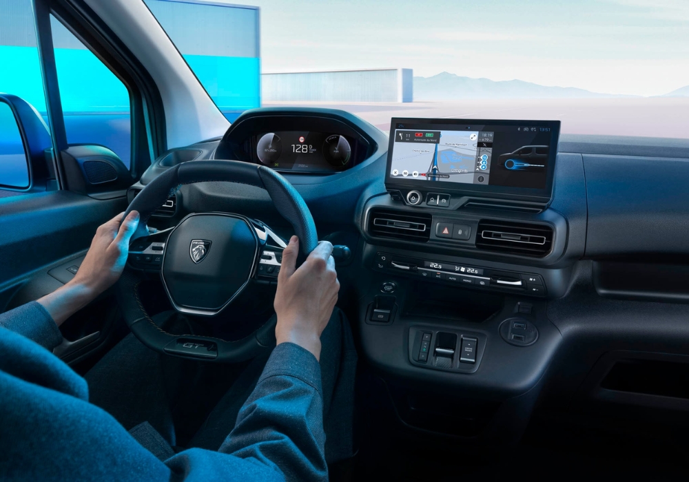 Peugeot e-Partner interior with driver holding steering wheel and digital display showing navigation and driving data.