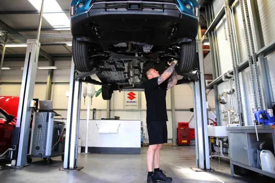 Technician working beneath a lifted vehicle inside a Suzuki service workshop.
