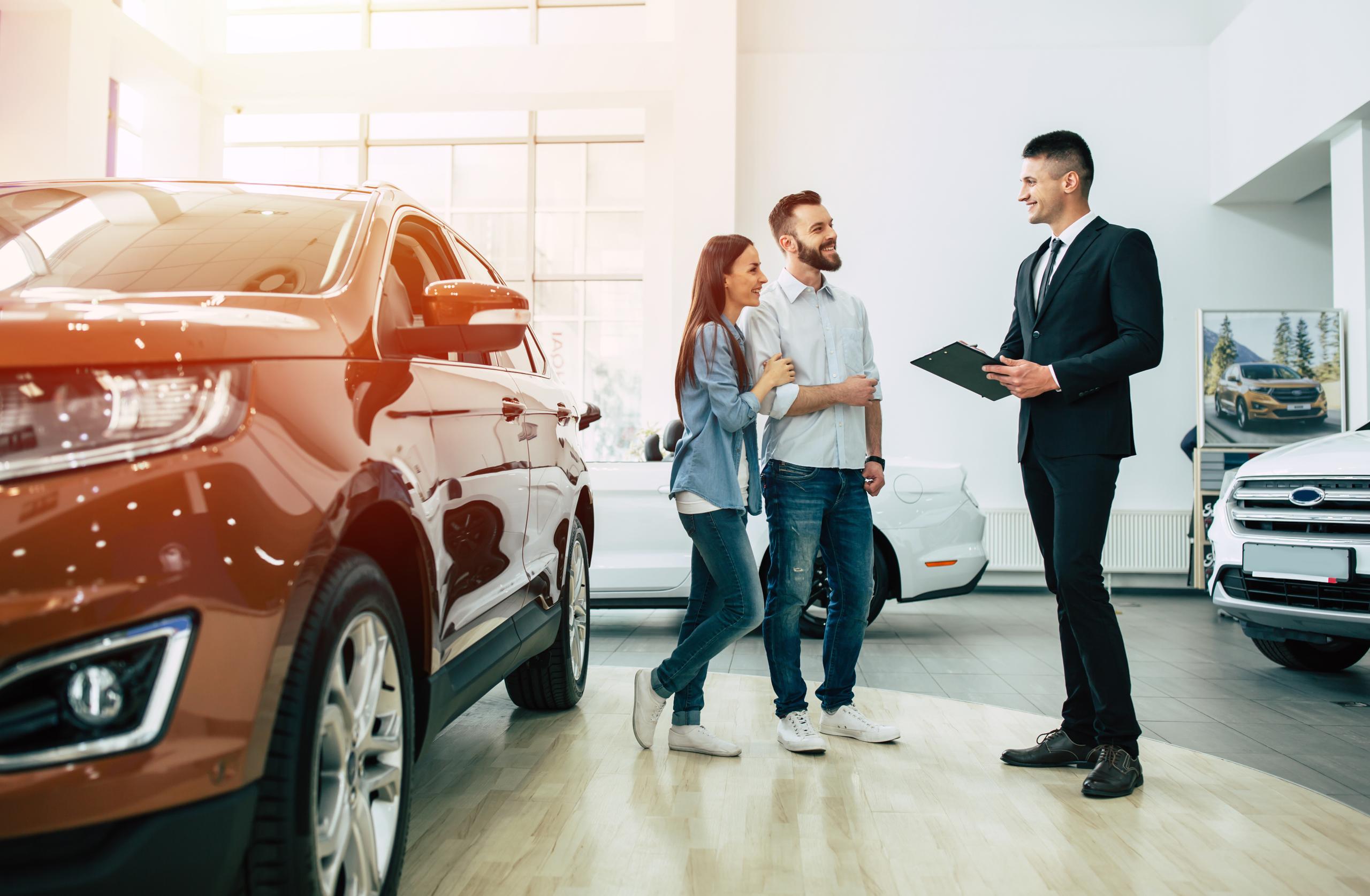 Couple speaking with a car salesperson inside a dealership showroom beside display vehicles.