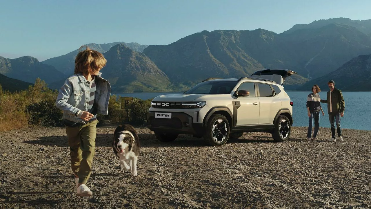 Dacia Duster parked by a lakeside with boot open, as a family walks nearby and a child runs with a dog in a mountainous setting.