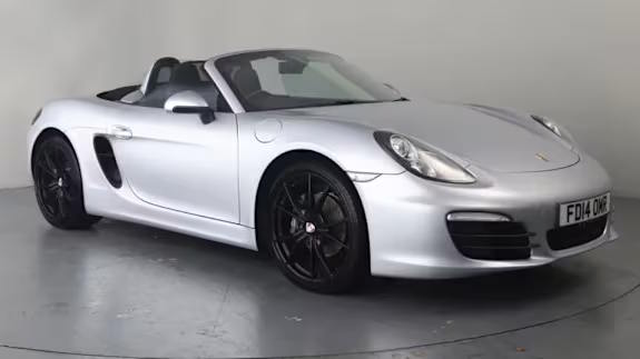 Silver Porsche Boxster convertible with black wheels, roof down, parked in a studio against a light grey backdrop.