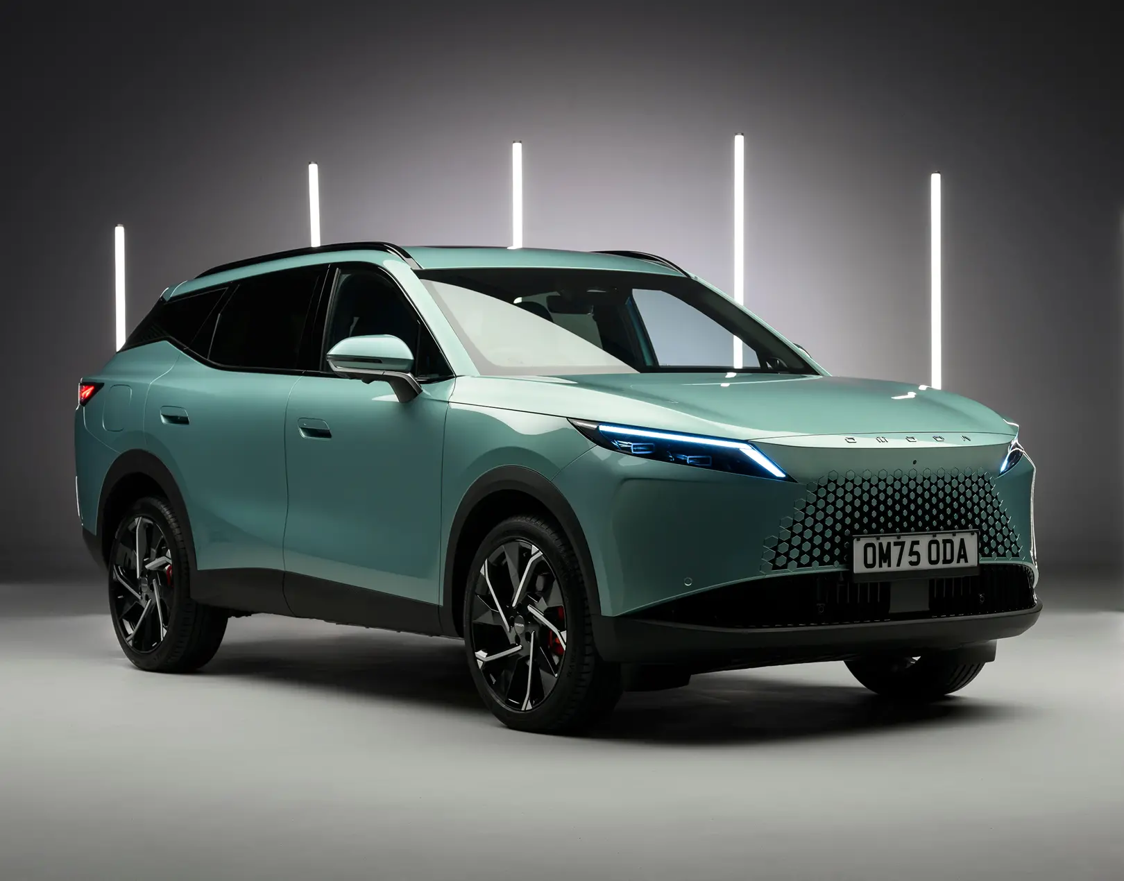 Teal Omoda E5 electric SUV in a studio setting with vertical light bars, shown in a three-quarter front view highlighting its closed grille and slim LED headlights.