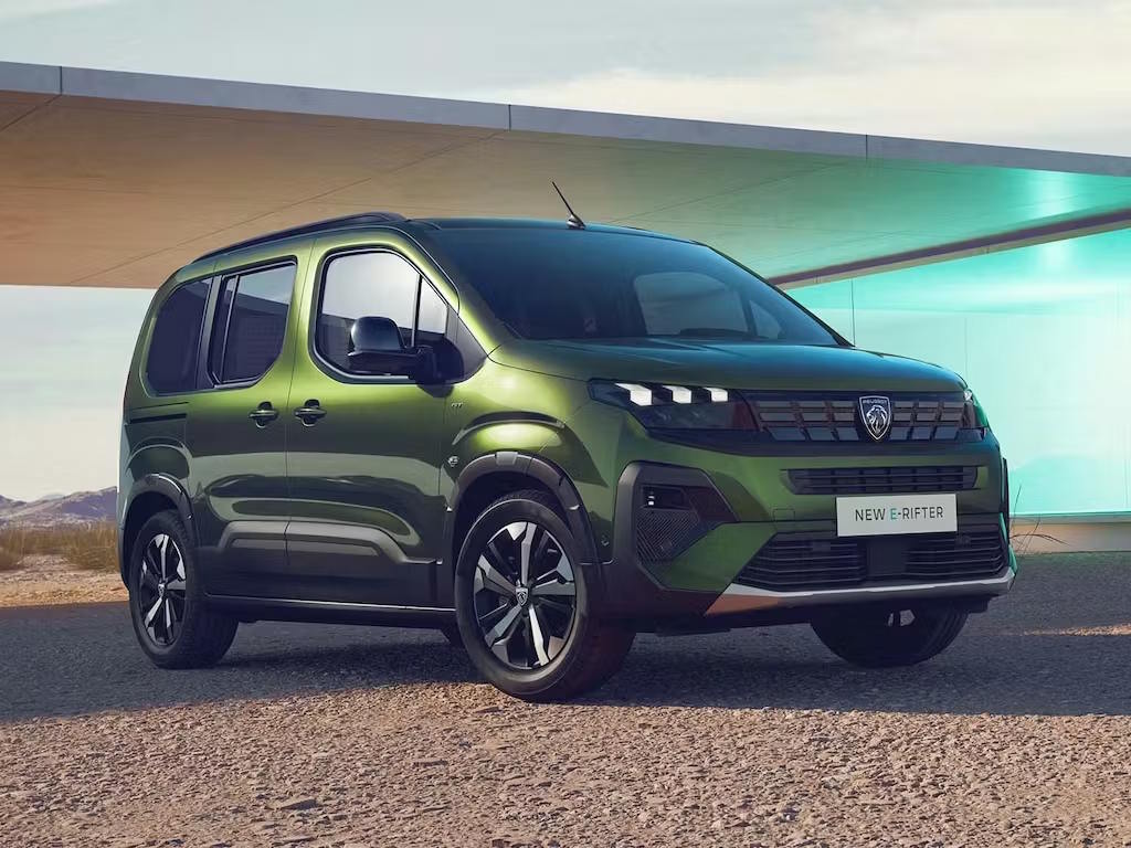 Front three-quarter view of the new Peugeot E-Rifter in metallic green, featuring the bold lion badge grille, distinctive LED daytime running lights and rugged MPV styling with black wheel arch cladding.