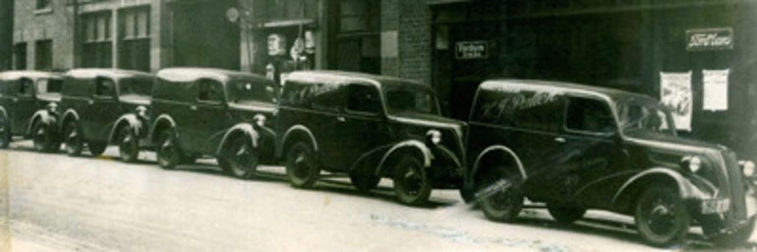 Row of vintage sedans parked along a street in front of commercial buildings, photographed in black and white.
