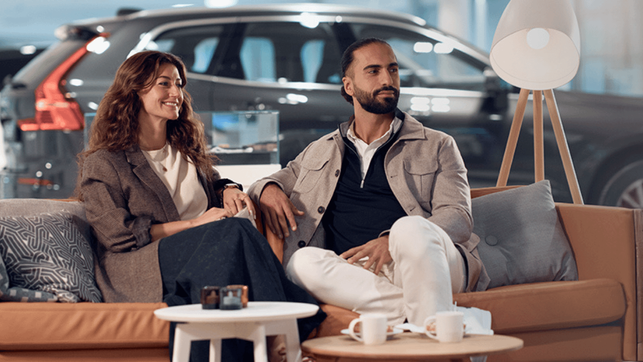 Customers relaxing in dealership lounge area beside Volvo vehicle.