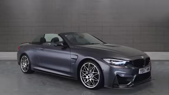 Dark grey BMW M4 convertible with multi‑spoke alloy wheels, roof down, parked in a studio with a tiled grey wall.