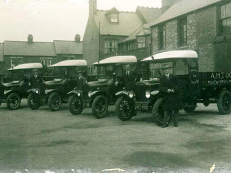 Early 20th-century delivery vans lined up with drivers standing beside them in front of a row of brick buildings.