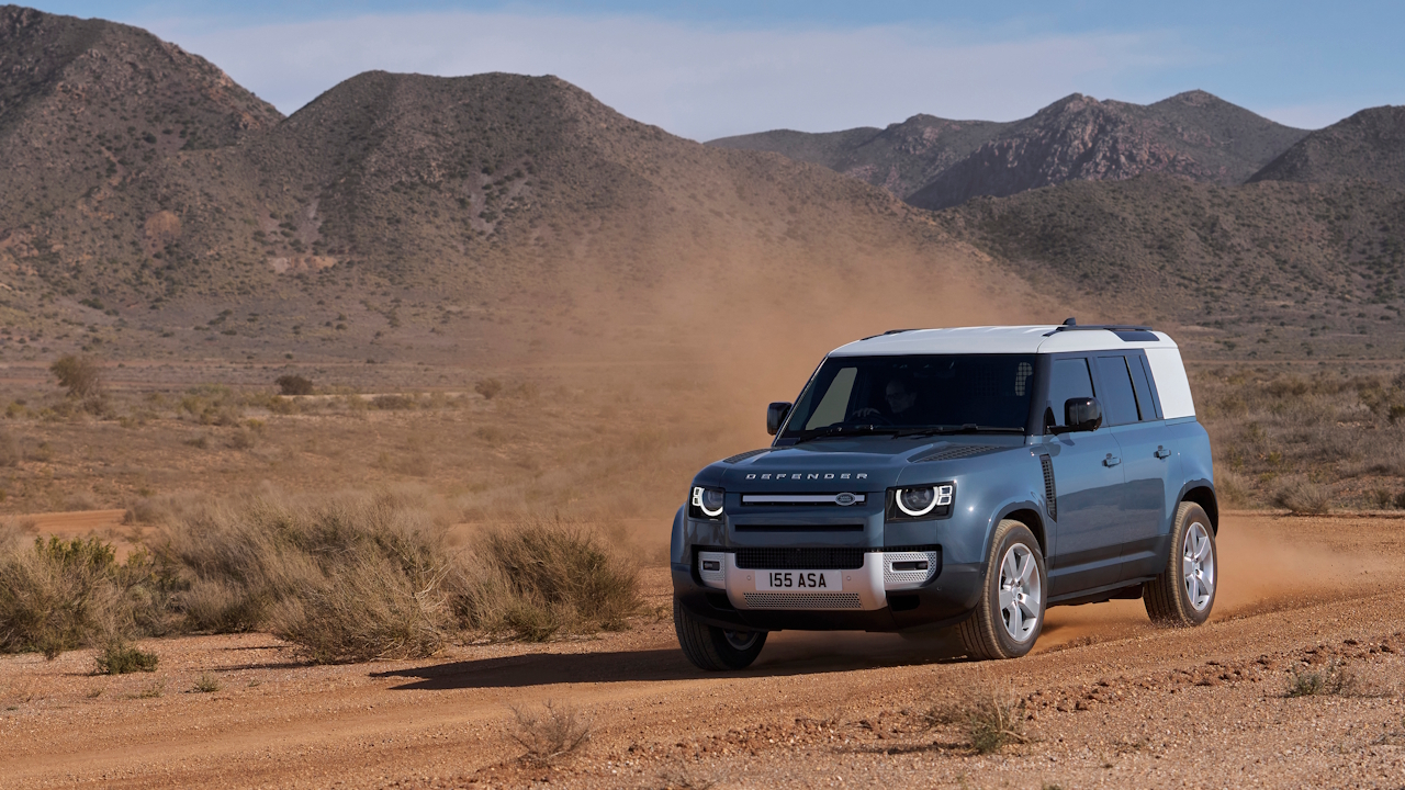 Front three-quarter view of the Defender Hard Top kicking up dust while driving on an off-road track with mountains in the background.