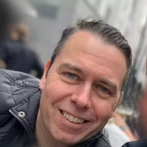 Close-up portrait of a smiling person wearing a quilted jacket in an urban setting.
