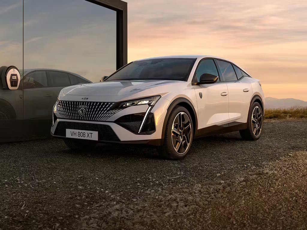 Front three-quarter view of the Peugeot E-408 electric fastback in pearl white, featuring signature fang-style LED daytime running lights, bold frameless grille and sculpted multi-spoke alloy wheels.