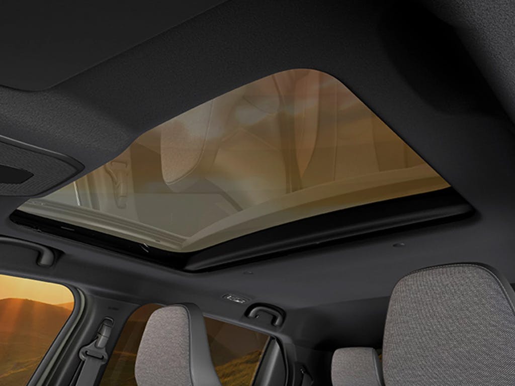 Interior view of the Suzuki e Vitara showcasing the expansive panoramic glass sunroof, dark headlining and light grey cloth seat upholstery with warm sunset light filtering through.