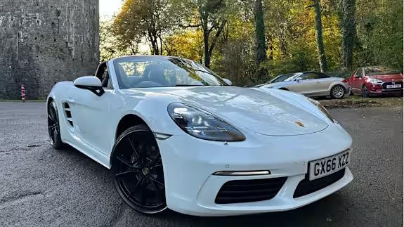 White Porsche 718 Boxster convertible with black wheels, roof down, parked on a road near a stone tower and trees, viewed from a low front three‑quarter angle.