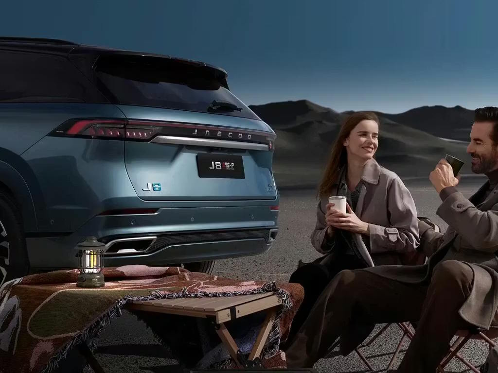 Rear view of the Jaecoo 8 SUV in a lifestyle setting, featuring full-width LED tail lights, roof-mounted spoiler and chrome exhaust detailing, with a couple enjoying a picnic beside it.