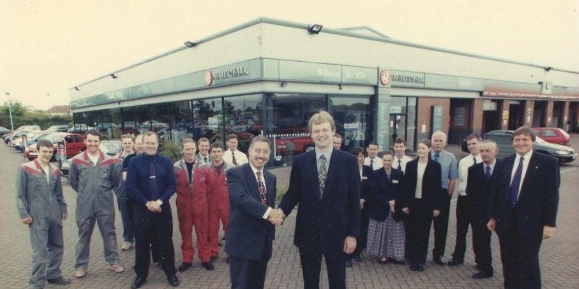 Historic Wilson and Co Vauxhall dealership staff photo with two men shaking hands outside the showroom.