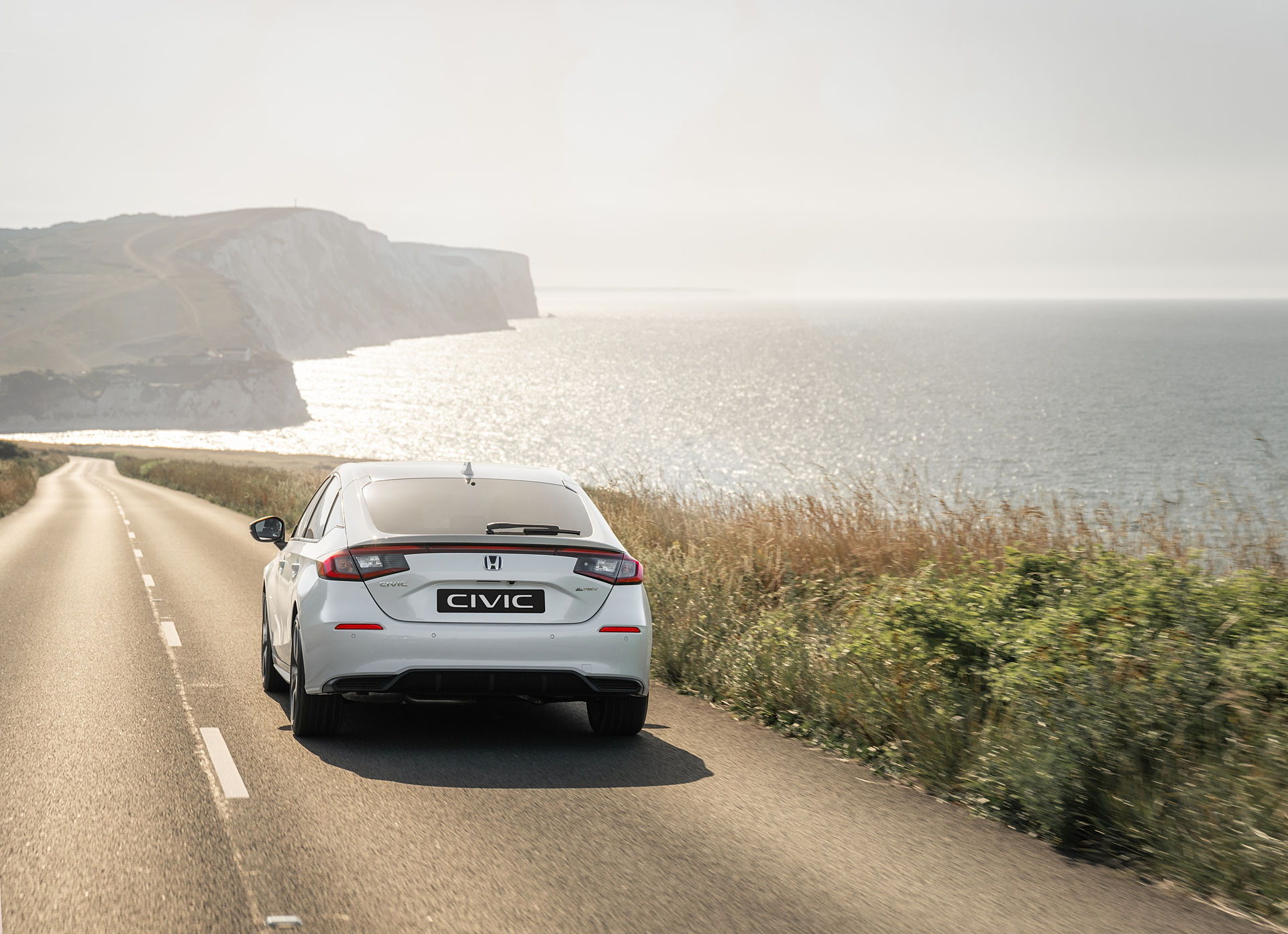 Silver Honda Civic hybrid driving up a hill with cliffs and water in the background