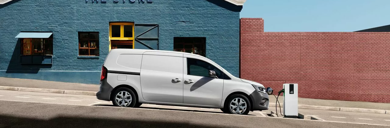 Profile view of a white Renault Kangoo E-Tech electric van charging at a pavement station in front of a blue brick building.