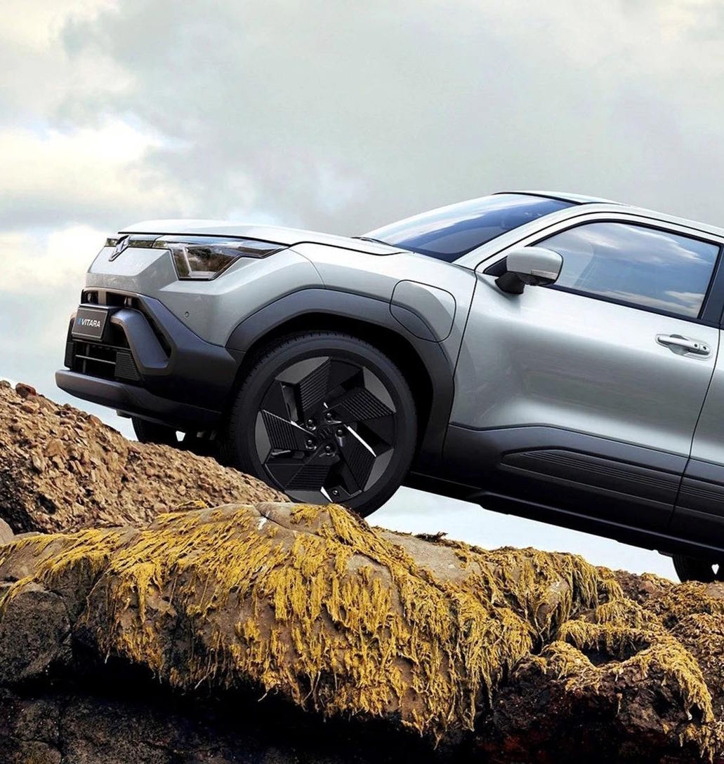 Front three-quarter view of the Suzuki e Vitara compact SUV in silver, conquering rocky terrain with bold black wheel arch cladding and striking angular LED headlights.