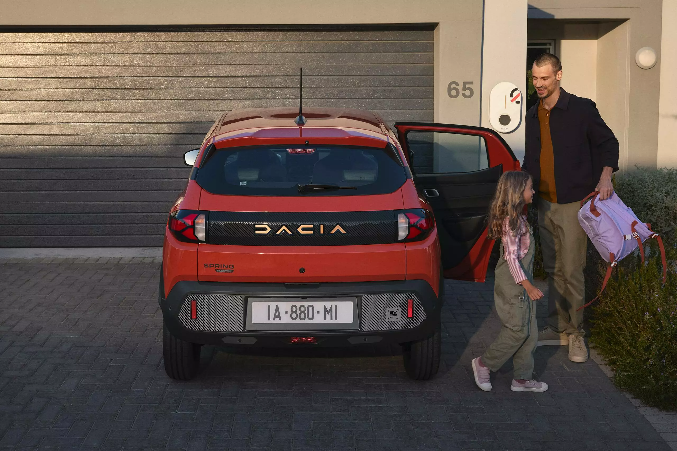 Dacia Spring parked outside a home while a man and child step out, with a charging point mounted on the wall beside the driveway.