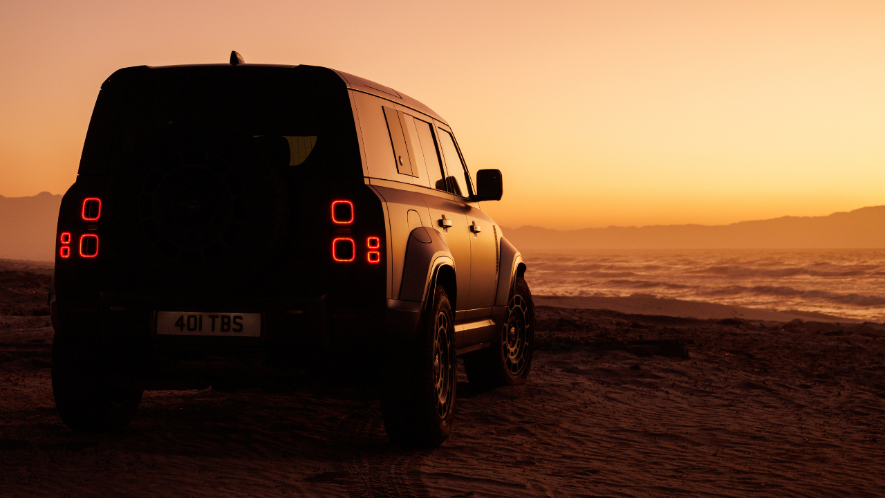 Rear view of a Land Rover Defender parked on desert terrain at sunset, overlooking a vast landscape.