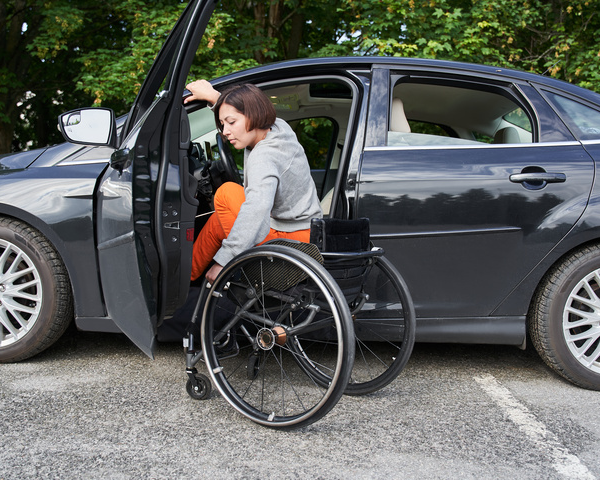 Wheelchair user transferring into a car from the driver’s seat with door open on a parking area.