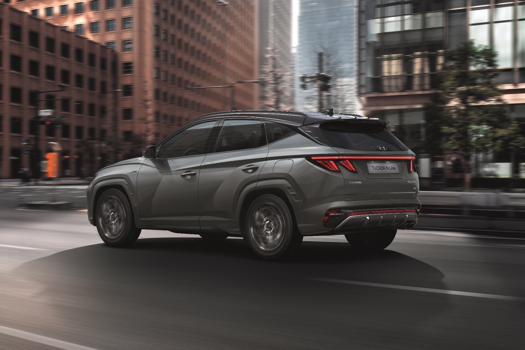 Rear-side view of a grey Hyundai Tucson SUV driving down a city street lined with tall buildings