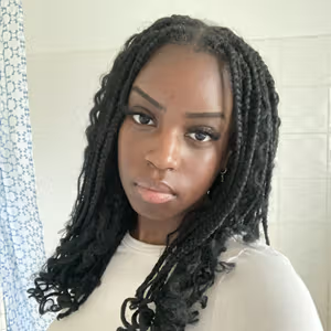 Portrait of a person with long braided hair wearing a light-coloured top indoors.