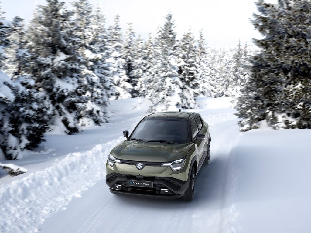 Front three-quarter view of the Suzuki e-Vitara compact SUV in khaki green, navigating a snow-covered mountain road surrounded by frost-laden pine trees.