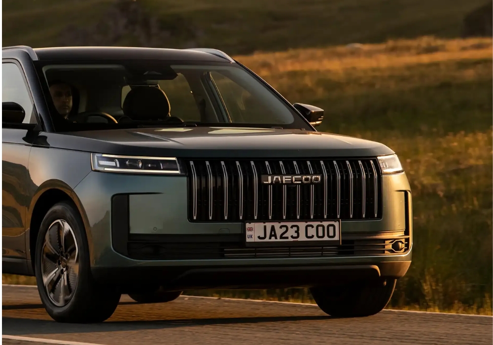 Front three-quarter close-up of a green Jaecoo 5 driving on a road at sunset, highlighting the grille and headlights.