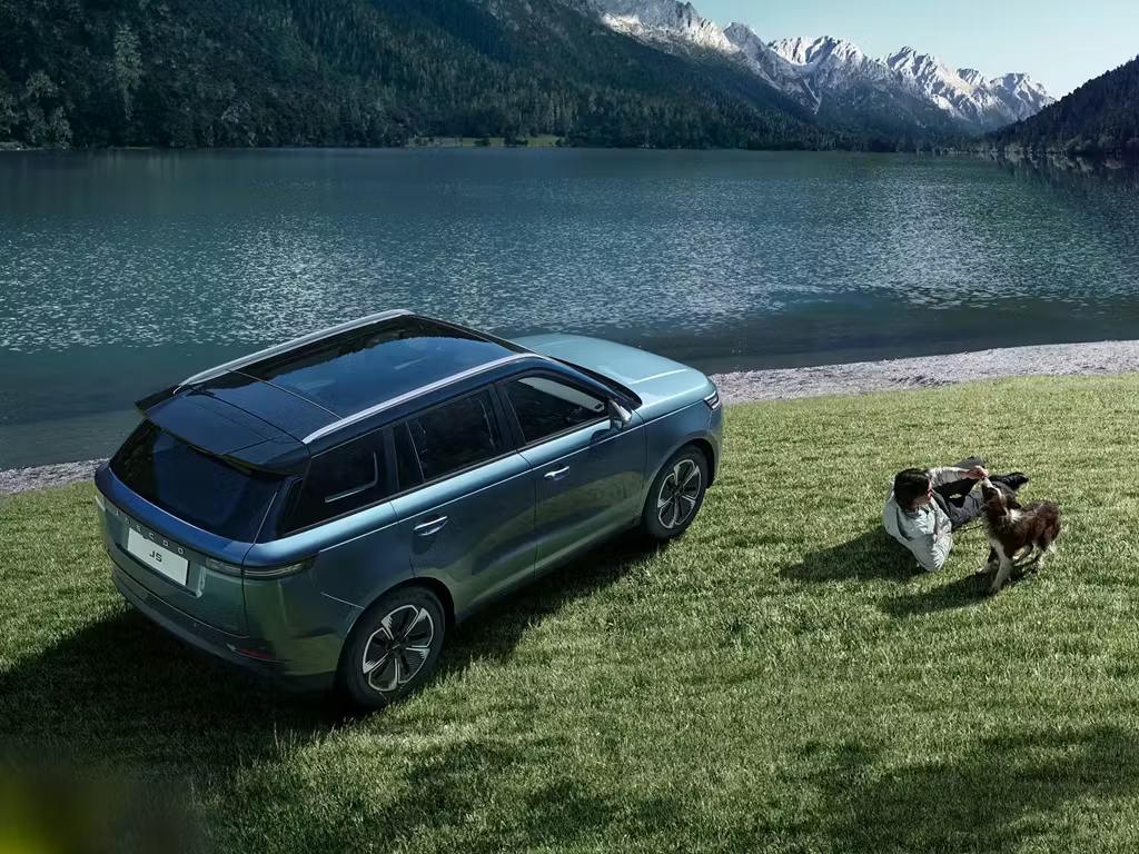 Elevated rear three-quarter view of the Jaecoo 5 SUV in metallic blue, parked lakeside with panoramic roof, sculpted bodywork and multi-spoke alloy wheels on display.