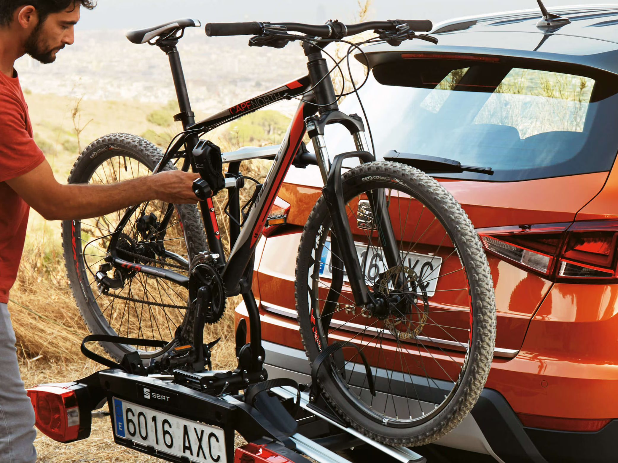 Man securing a mountain bike onto a rear-mounted bike rack attached to an orange SEAT Arona parked in a rural setting.