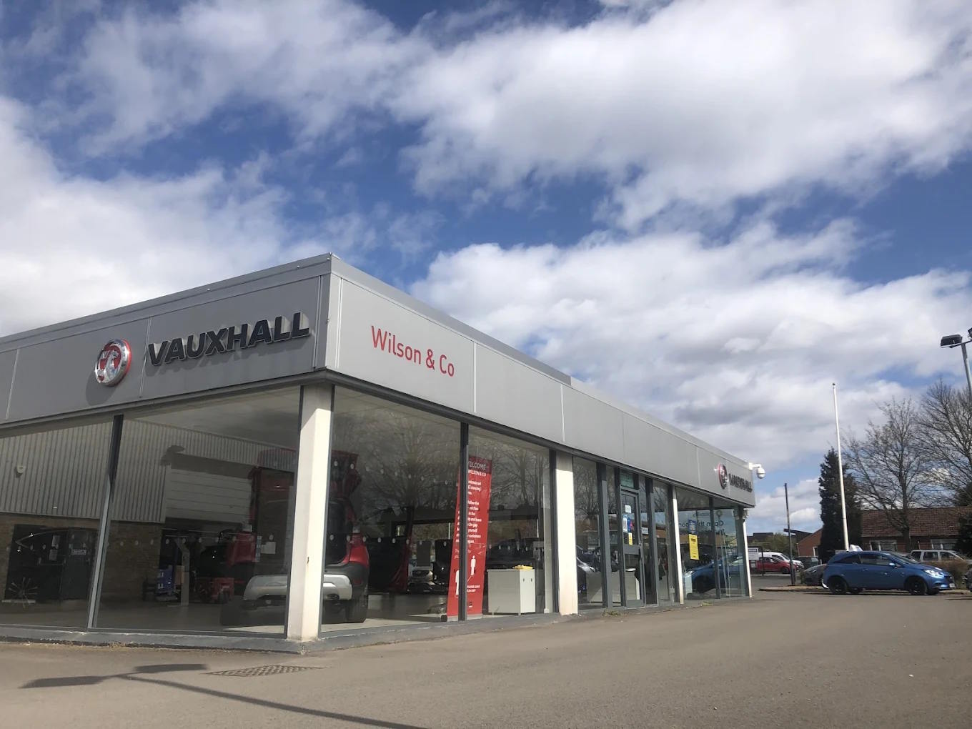 Exterior view of Wilson & Co Vauxhall Scunthorpe dealership, featuring a modern glass-fronted showroom with Vauxhall branding and vehicles on display under a bright blue sky.